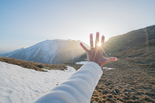 Pov of human hand stretching towards the sun at sunset. Sunbeam effect, mountains on background. Energy people environment concept.