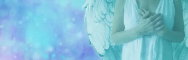 Cropped Angel showing torso in white robes with hands held over heart on a misty blue bokeh background with copy space on left hand side