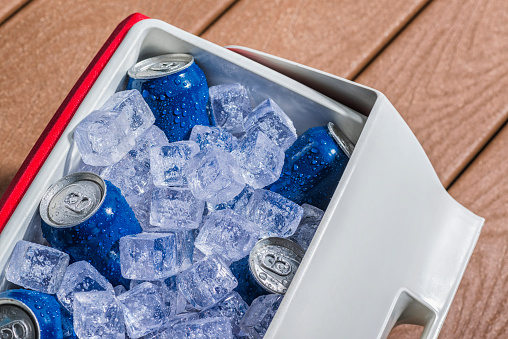 Looking down on a red and white personal cooler with a six pack of blue aluminum cans on ice sitting on a deck outside. These cans can be either soda, beer or other beverage.