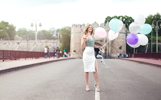 Beautiful girl is standing in the middle of the road on the bridge, holding a bunch of balloons. She is celebrating her 18th birthday and she is on the way to a party.