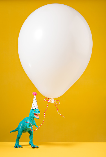 Teal dinosaur toy with birthday hat holding a white balloon on a yellow background.