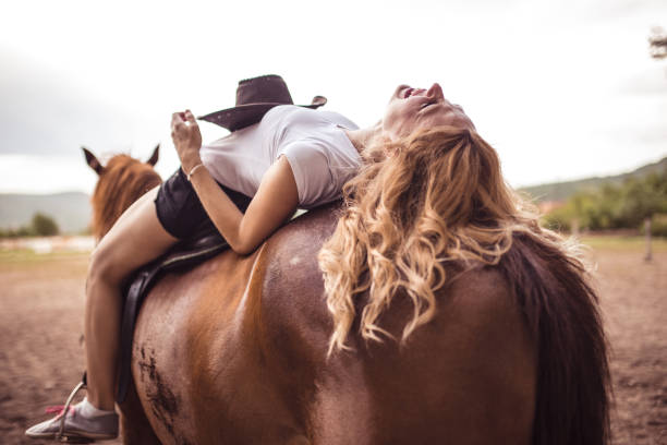Happy Asian woman sitting on a fence feeding a carrot to a white horse at a farm during sunset.