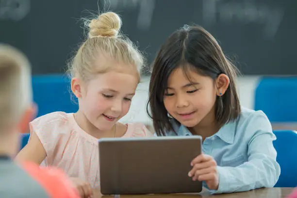 A multi-ethnic group of elementary age school children look at a digital tablet together. Young blonde Caucasian girl is wearing a pink top and young Asian brunette girl is wearing a blue button-up blouse.