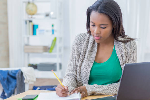 African American teenager concentrates while working on homework assignment Beautiful African American college student works on assignment in her dorm room. She is writing something in a notebook. A laptop is on her desk. female high school student stock pictures, royalty-free photos & images