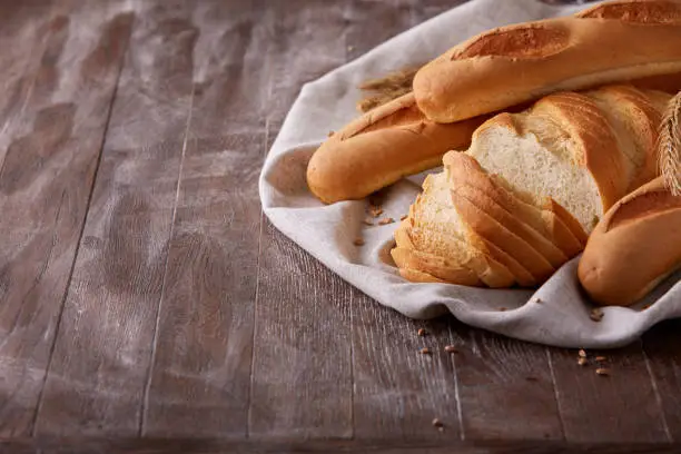 Slices of white bread and baguette laying on white cloth with gradient dark-brown background. Wooden table. With flour and wheat. Delicious. Tasty food.