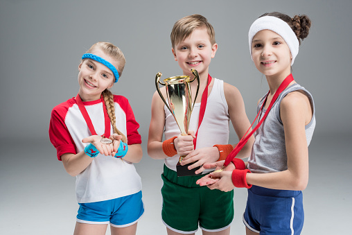 smiling boy and girls with medals and champion goblet isolated on grey