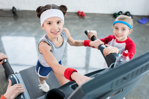 Adorable little girls in sportswear exercising on treadmill and smiling at camera in gym, children sport school concept