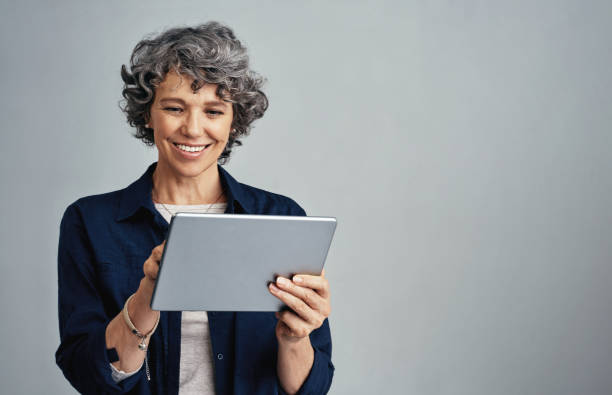 Managing daily life the smart way Studio shot of a mature woman using a digital tablet against a gray background old woman smiling tablet stock pictures, royalty-free photos & images