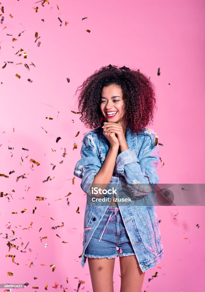 Portrait of happy young afro woman among confetti Studio portrait of surprised young afro woman wearing denim shorts and jacket, standing among confetti against pink background. Confetti Stock Photo