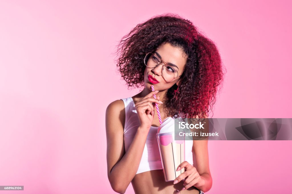 Cute young afro woman holding drink Studio portrait of cute young afro woman holding drink. Pink background. Smirking Stock Photo