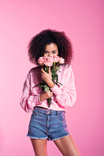 Studio portrait of young afro woman wearing denim shorts holding bunch of pink roses. Pink background.