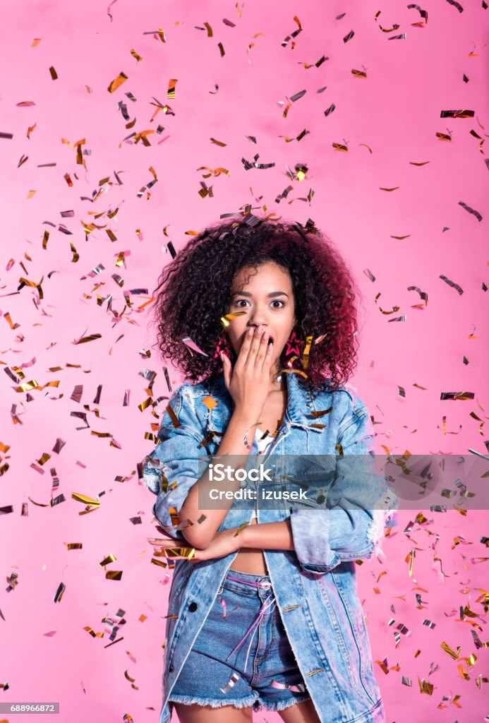 Portrait of surprised young afro woman among confetti Studio portrait of surprised young afro woman wearing denim shorts and jacket, standing among confetti against pink background, raising hands. Women Stock Photo