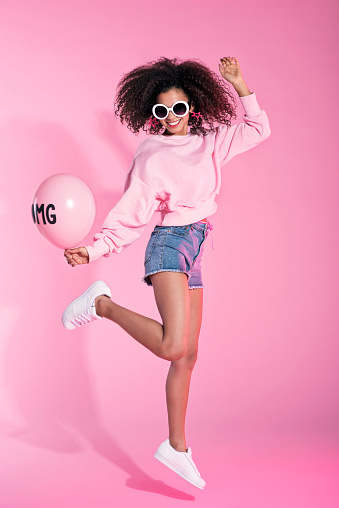 Studio portrait of young afro woman wearing denim shorts and sunglasses, holding balloon and jumping. Pink background.
