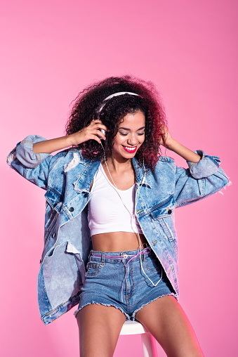 Studio portrait of young afro woman wearing denim shorts and jacket listening to music. Pink background.