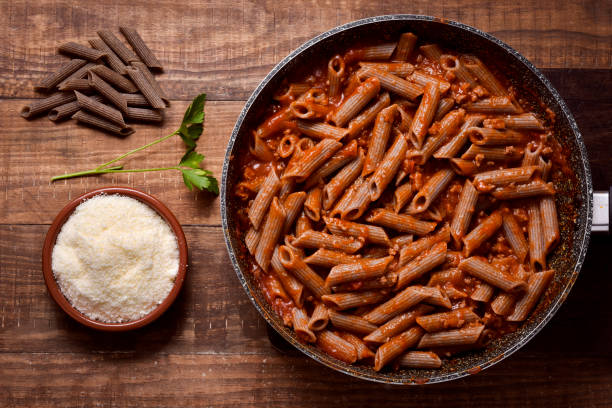 buckwheat penne rigate bolognese high-angle shot of a frying pan with buckwheat penne rigate alla bolognese, a bunch of uncooked buckwheat penne rigate and an earthenware bowl with grated Parmigiano cheese, on a rustic wooden table whole wheat stock pictures, royalty-free photos & images