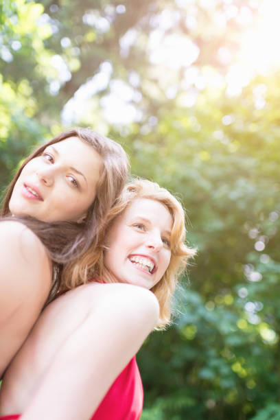 Young woman having fun, sitting back to back on meadow enjoying their summer vacation, laughing, smiling. Women Having Fun Summer Lifestyle Vacation Series.