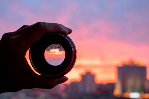 Photo of hand holding camera lens, looking at scenics of glowing clouds at sunset