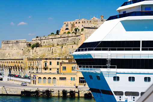 Valletta, Malta: A white tourist cruise ship sit in Valletta’s harbor in the foreground, with the fortified city walls in the background. Copy space available in blue sky.