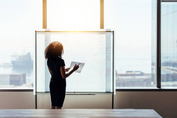 Businesswoman making a business plan presentation in office. Rear view of a businesswoman writing on a whiteboard at office. Business investor holding papers and pen standing in front of a presentation board. whiteboard visual aid stock pictures, royalty-free photos & images