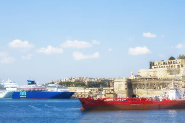Valletta, Malta: A blue tourist cruise ship and a red tanker sit in Valletta’s pretty Grand Harbor, with the fortified city walls and Upper Barrakka Gardens visible in the background. Copy space available in the sea or blue sky that is dotted with puffy white clouds.