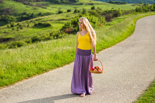 Beautiful woman walking at the country road and holding basket full of apples.