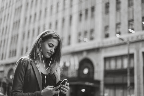 One woman, standing outdoors on the city street alone, using smart phone, listening music on headphones, black and white.