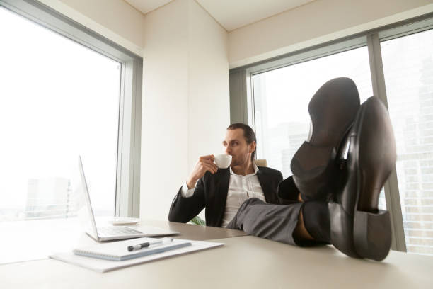 istock Businessman with legs on desk drinks coffee 689425558