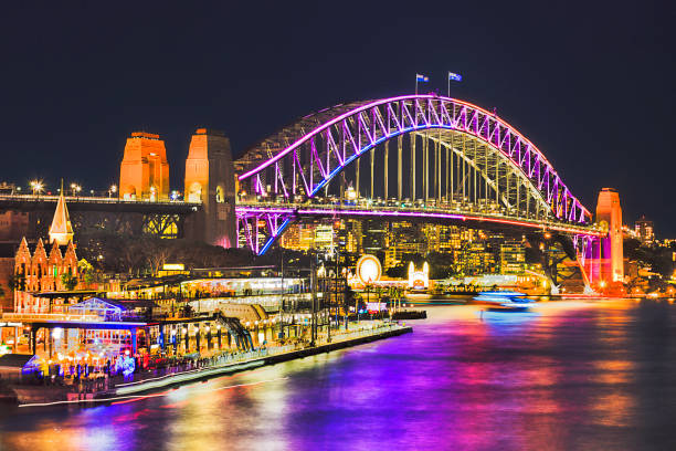 Sy vivid 2016 Br Fr Quay Close Huge arch of Sydney Harbour Bridge brightly illuminated during annual light show Sydney vivid sydney festival. Overseas passengers terminal and bridge reflect in blurred harbour waters. sydney harbour bridge stock pictures, royalty-free photos & images