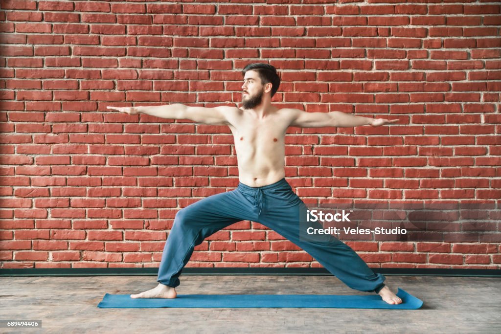 Attractive man doing yoga Handsome man with dark hair and beard wearing trousers doing yoga warrior position on blue matt at wall background, copy space, portrait, virabhadrasana. Abdomen Stock Photo