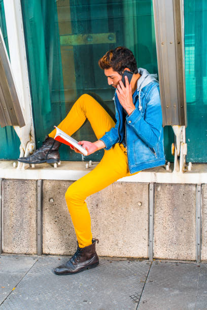 istock Young man reading book, talking on cell phone outside in New York 689507762
