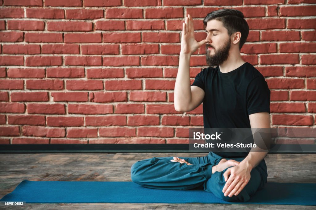 Handsome man doing yoga positions Handsome young man with a beard wearing black T-shirt doing yoga position on blue matt at wall background, copy space, portrait, pranayama exercises. Abdomen Stock Photo