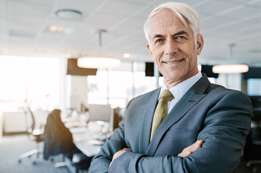 Portrait of successful senior businessman standing in office with his arms crossed. Caucasian male entrepreneur in suit looking at camera with a smile.
