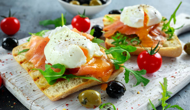 pochiertes ei auf gegrilltes toast mit geräuchertem lachs, rucola, oliven und gemüse auf weißen tafel. gesundes frühstück - poached stock-fotos und bilder