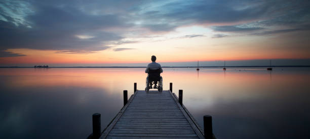 Photo of Senior man sitting in wheelchair at the end of lakeside jetty watching majestic cloudscape at dusk