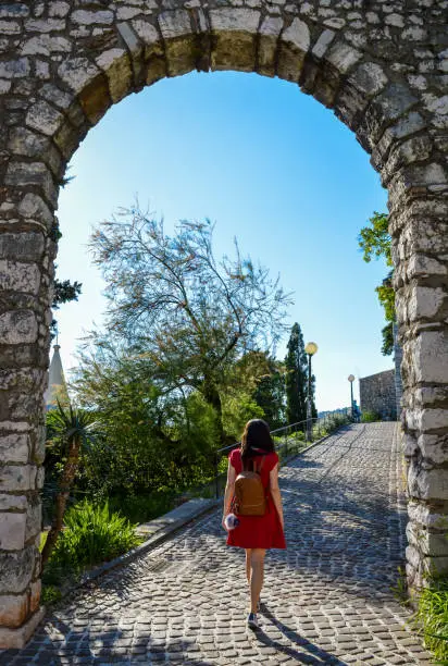 A young woman in a red summer dress, carrying a backpack, walking through the ancient, stone arch door, Rijeka, Croatia