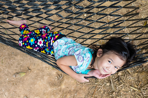 Top view of cheerful child enjoying and relaxing in hammock, outdoor at the daytime. Pretty asian girl smiling and looking at camera. Summer vacation or happy time concept.