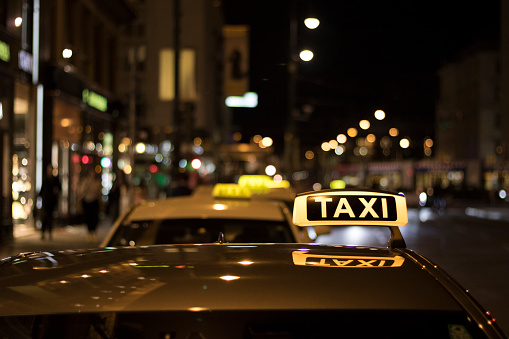 Close up of illuminated taxi sign on roof of a taxi waiting for passengers at night in Vienna showing other taxis on the stand and road with buildings in background.