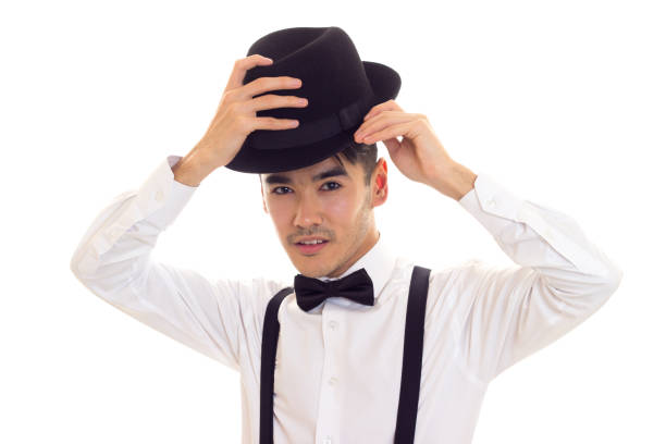 Smart-looking young man in white shirt with black bow-tie, suspenders and hat on white background in studio