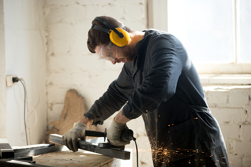 Worker in workwear, protective glasses, hearing protection headphones, gloves using dangerous electric power tools in loft workshop. Safety at workers workplace, personal protective equipment concept