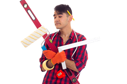 Young pleasant man with dark hair in red plaid shirt with orange gloves holding level, tape-measure, white roll and ruler on white background in studio