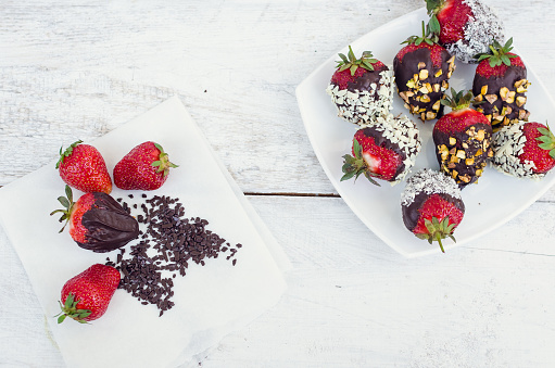 Variety of strawberries covered with a dark chocolate with nuts pistachios and coconut on the white wooden table. Homemade choco dipped berry. Gourmet summer dessert. Top view.