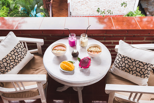 Healthy tropical breakfast on outdoor terrace at home. White vintage round table served with cutted mango and dragon fruit, fresh papaya smoothie and cold shake drinks in bottles