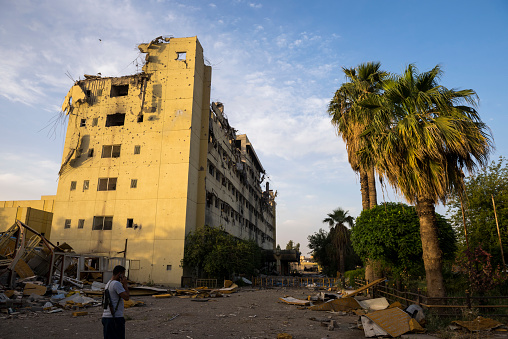 The landscape of Mosul, Iraq, is filled with destroyed and scarred buildings such as this one at Al-Salam hospital, a consequence of the Iraqi government, with U.S. support, wresting control of the city from ISIS. An armed man stands in the foreground.