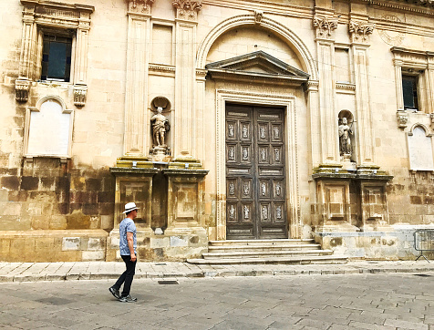 Chiaramonte (Ragusa Province), Sicily: An active senior male tourist in a white hat walking past the baroque Santa Maria La Nova Church, dating from 1609. Chiaramonte is a village in Ragusa Province.