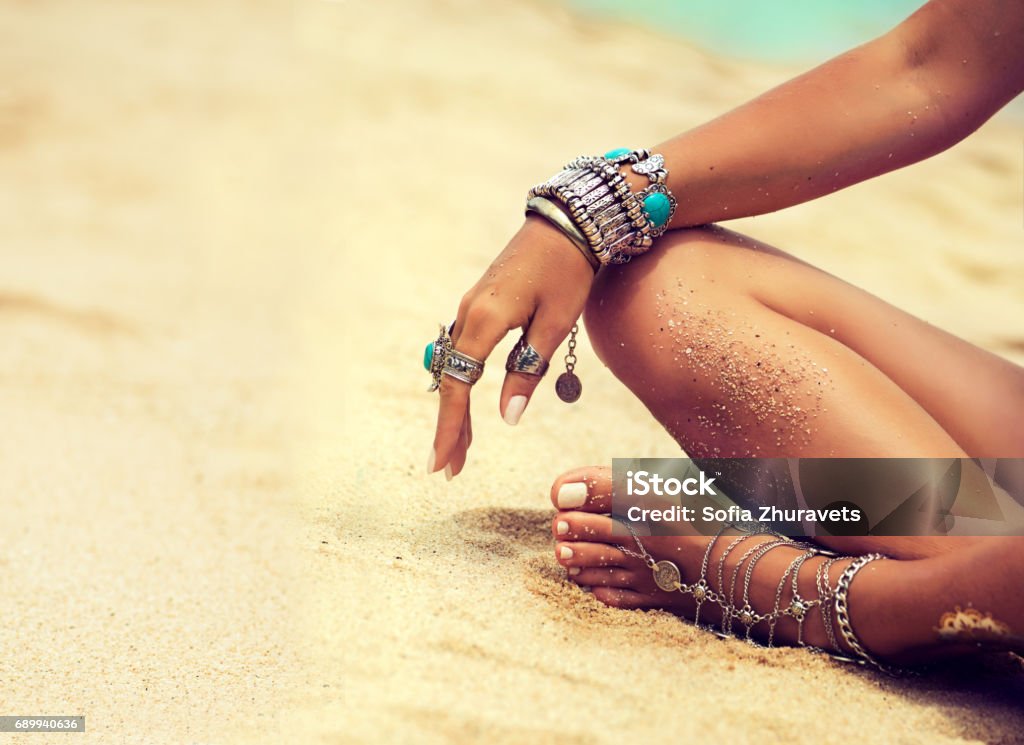 Tanned girl in lotus position dressed in silver jewelry,bracelets and rings.Boho style. Woman is sitting in relaxed position on tropical sandy beach. Body parts. Tanned girl in lotus position dressed in silver jewelry,bracelets and rings.Boho style. Jewelry Stock Photo