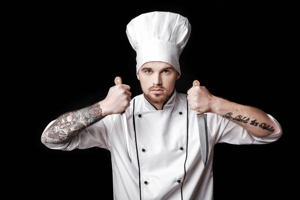 istock Young bearded man chef In white uniform holds Two knives on  black background 689948628