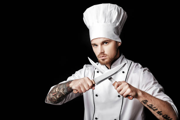 istock Young bearded man chef In white uniform holds Two knives on  black background 689948678