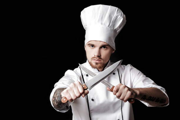 istock Young bearded man chef In white uniform holds Two knives on  black background. 
Focus on knives 689950910