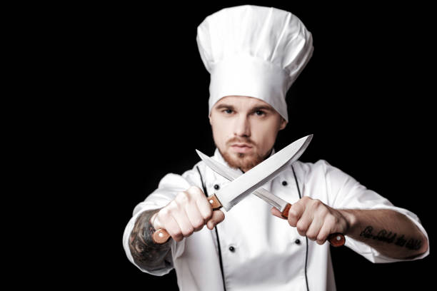 istock Young bearded man chef In white uniform holds Two knives on  black background. Focus on knives 689950944