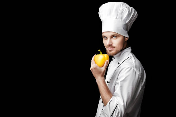 istock Young bearded man chef In white uniform holds yellow bell pepper on  black background 689952324